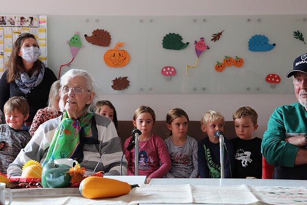 Ältere Frau sitzt am Tisch, im Hintergrund sitzt eine Kindergartengruppe