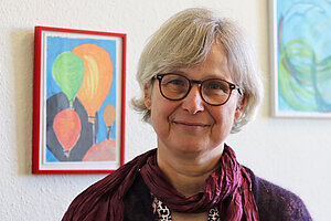 Eine Frau mit hellen halblangen Haaren und Brille mit dunklem Gestell steht vor einer weißen Wand, an der zwei bunte Bilder hängen.