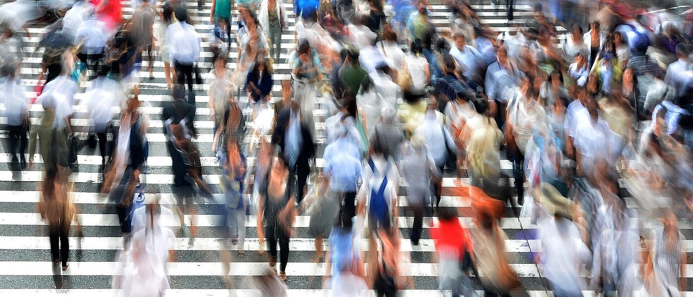 über einen sehr breiten Zebrastreifen laufen sehr viele Menschen. Sie sind nur unscharf abgebildet.