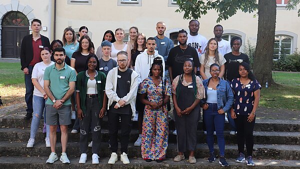 26 junge Frauen und Männer stehen im Freien in mehreren Reihen aufgereiht auf einer Treppe und lachen in die Kamera. 