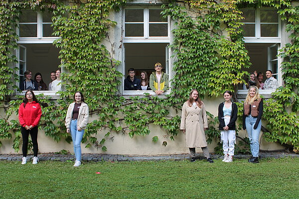 Gruppenaufnahme mit 15 jungen Personen. Zehn Personen schauen aus drei Fenster, fünf Mädchen stehen auf dem Rasen vor dem Gebäude.