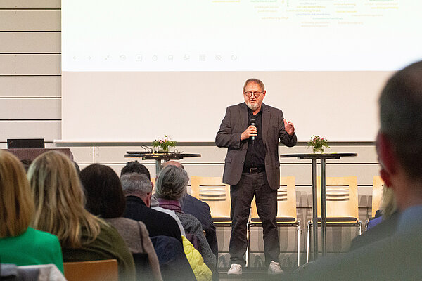 Ein Mann im Anzug steht auf einer niedrigen Bühne, hinter ihm eine Präsentationsleinwand, vor ihm zahlreiche von hinten zu sehende Zuhörer:innen.