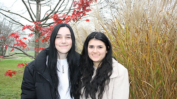Zwei junge Frauen stehen eng nebeneinander im Freien vor einem herbstlichen Hintergrund.
