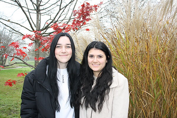 Zwei junge Frauen stehen eng nebeneinander im Freien vor einem herbstlichen Hintergrund.