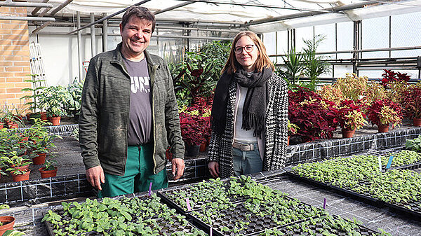 Ein Mann in Arbeitskleidung und eine junge Frau stehen lächelnd an langen Pflanztischen in einem Gewächshaus.