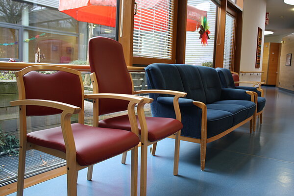 Man sieht einen breiten, roten Stuhl mit niedriger Lehne, einen roten, schmalen Stuhl mit hoher Lehne und einem blauen Sofa daneben in einem Flur vor einem Fenster stehen.