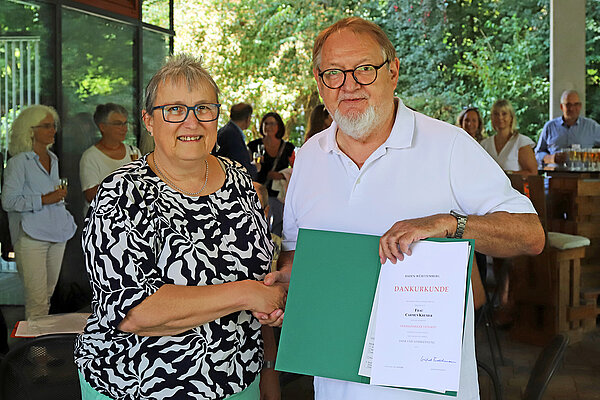 Eine Frau und ein Mann reichen sich die Hand und lächeln dabei in die Kamera, im Hintergrund sind die Gäste der kleinen Feierlichkeit im Freien zu erkennen.