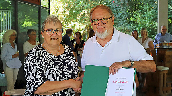 Eine Frau und ein Mann reichen sich die Hand und lächeln dabei in die Kamera, im Hintergrund sind die Gäste der kleinen Feierlichkeit im Freien zu erkennen.