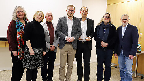 Drei Frauen und vier Männer stehen, festlich gekleidet in einem hellen Raum, zum Gruppenfoto beisammen und schauen lächelnd in die Kamera.