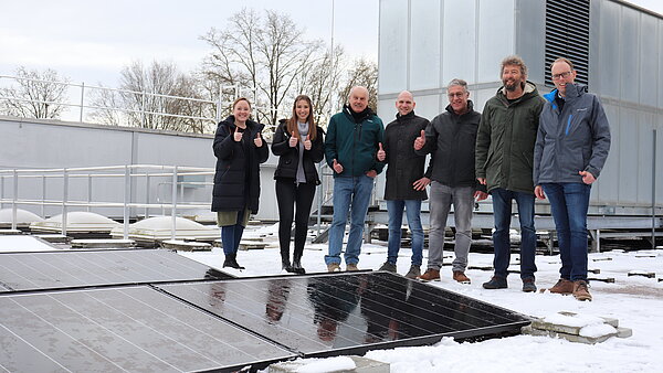 Zwei Frauen und fünf Männer stehen auf dem Flachdach eines Gebäudes. Im Vordergrund sind mehrere Solarpanels zu erkennen. 