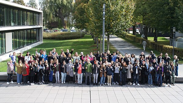 Auf einem gepflasterten Platz vor einem Gebäude stehen über 90 Personen in mehreren Reihen hinter- und nebeneinander. Sie schauen in die Kamera und strecken ihre Daumen nach oben. Im Hintergrund sind eine grüne Wiese und Bäume. 