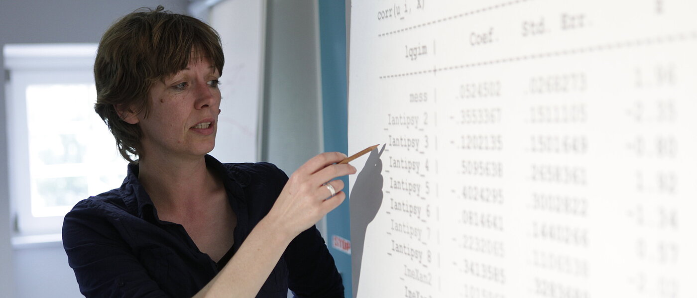 Eine Frau mittleren Alters mit kürzeren Haaren steht in einem Schulungsraum, in ihrer rechten Hand hält sie einen Bleistift, mit diesem zeigt sie auf eine von einem Overhead-Projektor an die weiße Wand projizierte Tabelle, welche diverse Code- und Zahlenreihen auflistet.