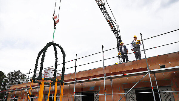 Zwei Männer in Handwerksmontur stehen auf dem Flachdach eines Gebäude-Rohbaus, vor ihnen hängt eine traditionelle Richtglocke am Haken eines Baukrans, der im Hintergrund steht.