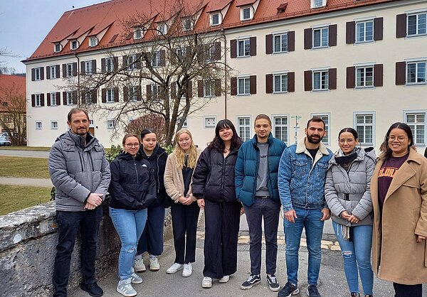 Eine Gruppe junger Menschen steht im Freien auf einer steinernen Brück, im Hintergrund ist ein helles Gebäude zu sehen. 