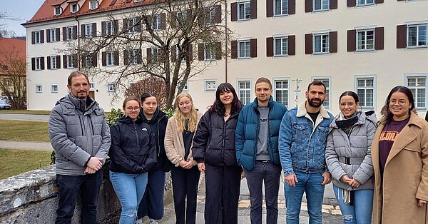 Eine Gruppe junger Menschen steht im Freien auf einer steinernen Brück, im Hintergrund ist ein helles Gebäude zu sehen.
