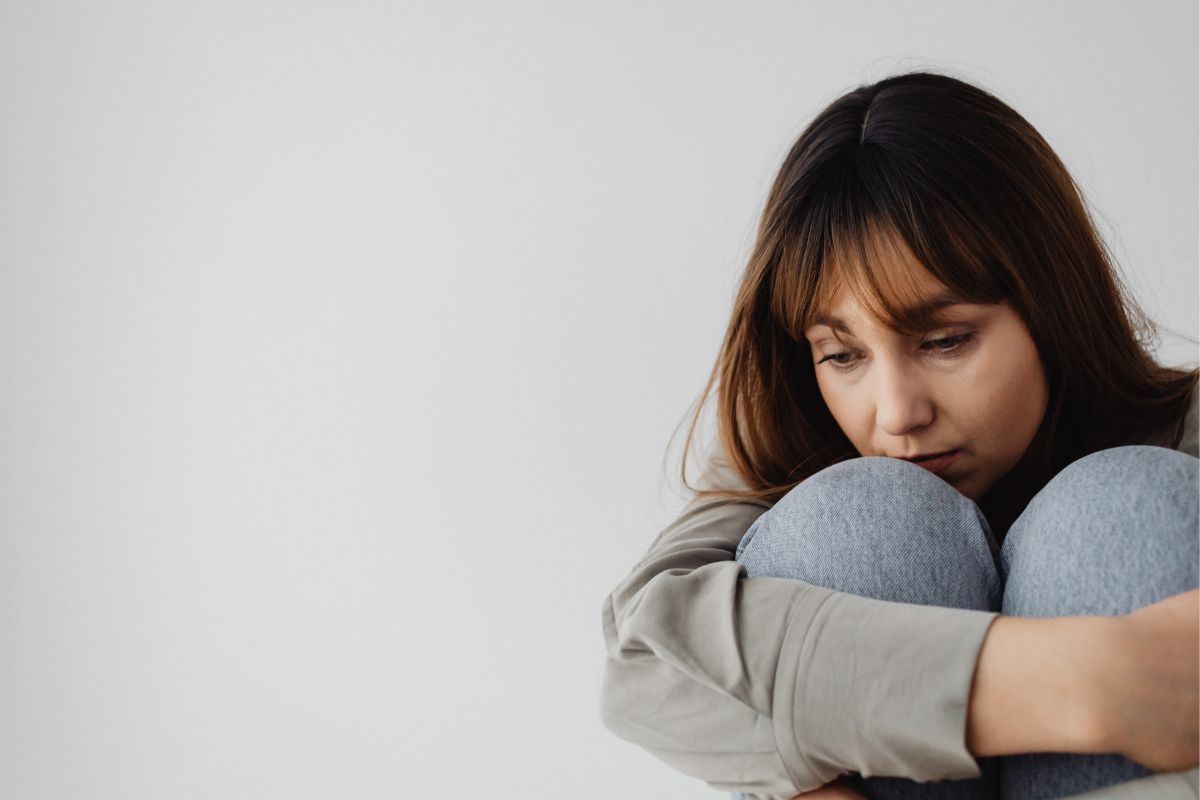 Eine junge dunkelhaarige Frau kauert am Boden und umschließt ihre Beine, ihr Blick geht Richtung Boden.