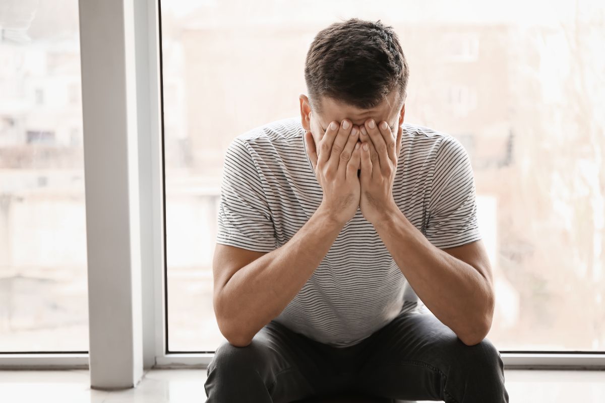 Ein Mann sitzt innen vor einem Fenster und hat die Hände in seinem Gesicht vergraben.