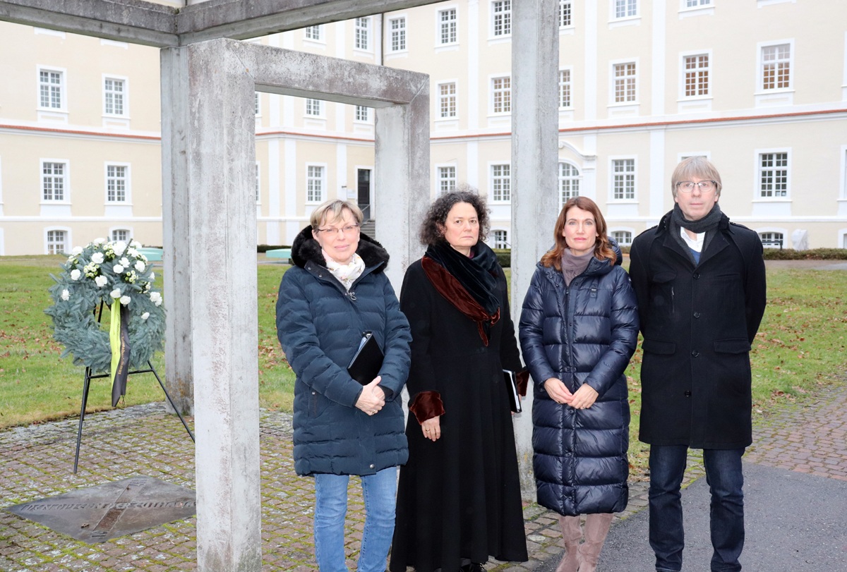 Vier Personen stehen mit ernstem Blick vor einem Mahnmal aus Beton, im Hintergrund ist ein Kranz zu sehen.