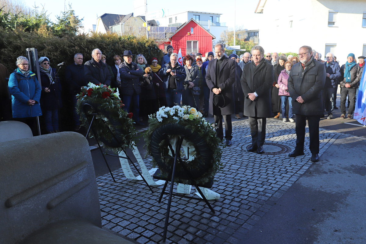 Drei Männer stehen in stillem Gedenken vor dem Denkmal der grauen Busse und zwei Gedenkkränzen, umringt von zahlreichen Anteilnehmenden.