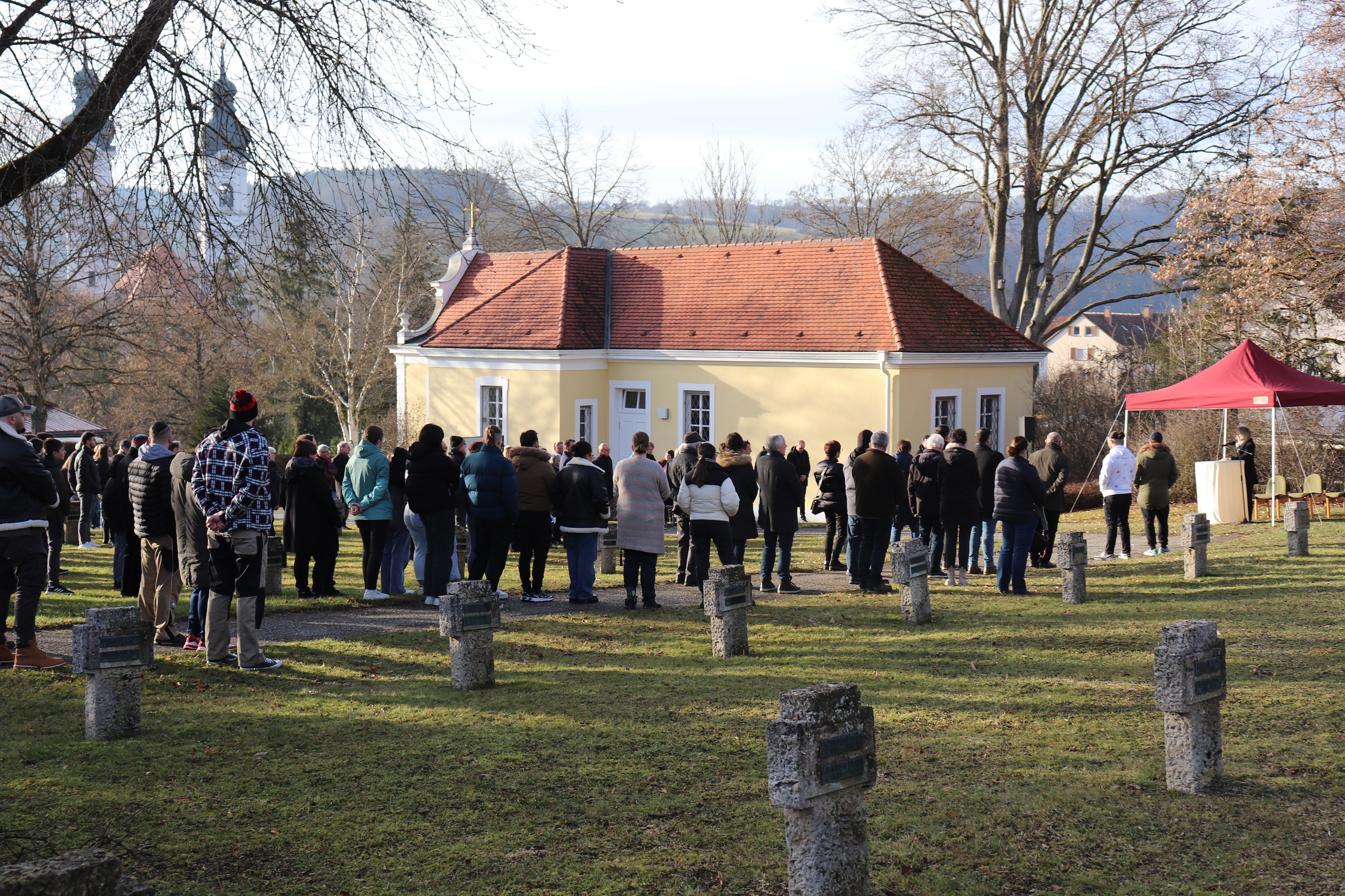 Eine große Gruppe Menschen steht auf einem Friedhof. Im Vordergrund sind einige steinerne Kreuze zu erkennen, im Hintergrund die Türme des Münsters. 