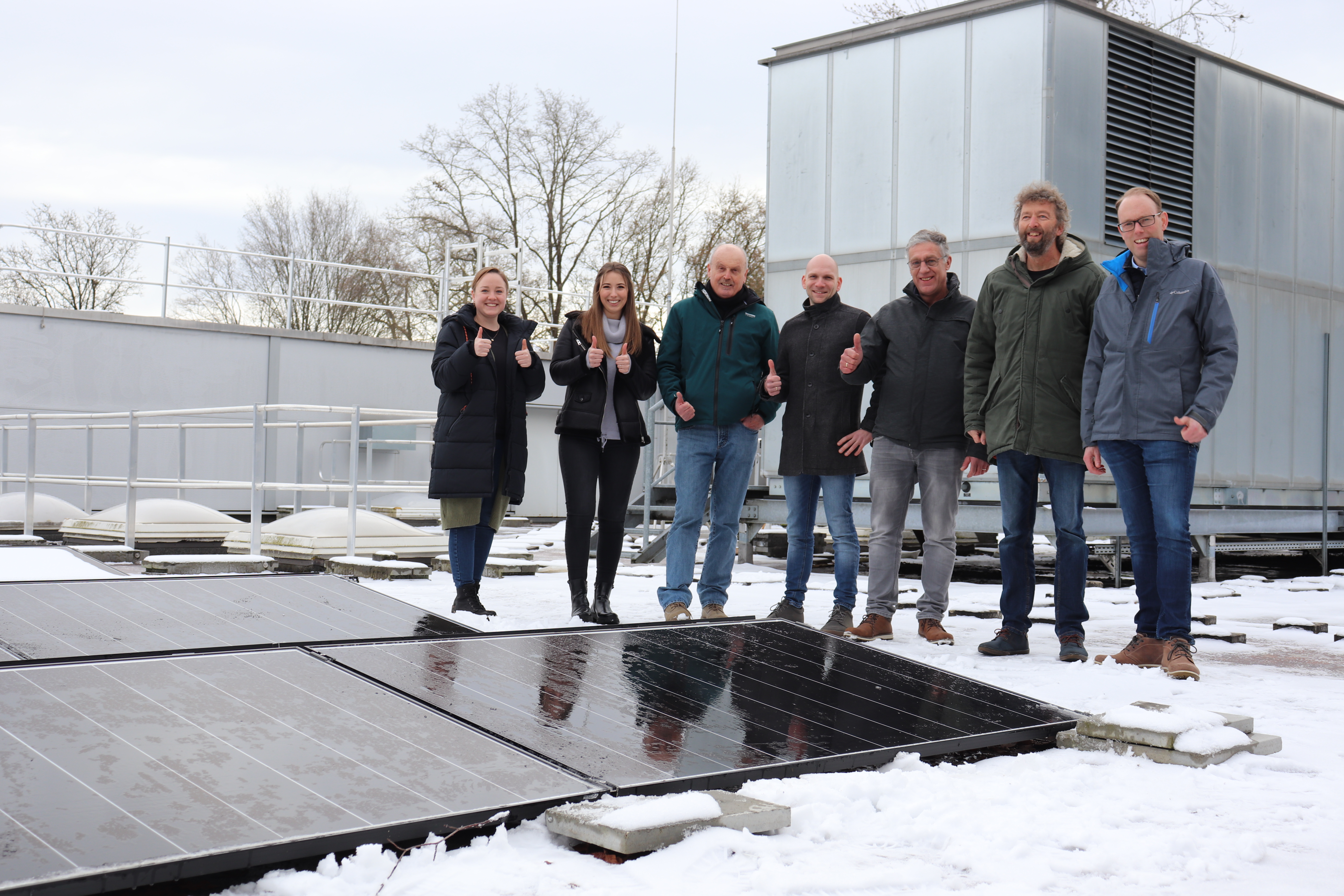 Zwei Frauen und fünf Männer stehen auf dem Flachdach eines Gebäudes. Im Vordergrund sind mehrere Solarpanels zu erkennen. 