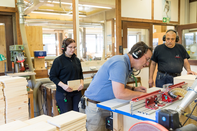 In einer kleinen Werkstatt bearbeitet ein Mann ein Holzstück an einer großen Säge. Zwei weitere Personen stehen daneben und schauen ihm zu.