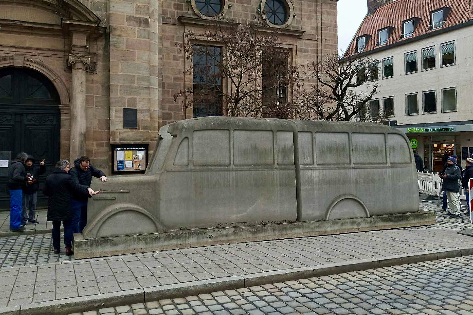 Das Denkmal der grauen Busse, eine mehrere Meter lange Nachbildung eines Omnibusses aus grauem Beton, steht auf gepflasterten Vorplatz einer Kirche. Links und rechts davon stehen Menschen.